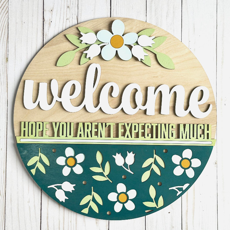 A round wooden welcome sign with floral patterns and the phrase 'Welcome Hope You Aren't Expecting Much' in a snarky style.
