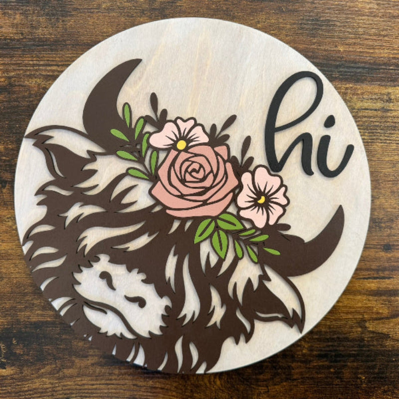 A round wooden welcome sign featuring a highland cow design with floral elements and the word 'hi' in the center.