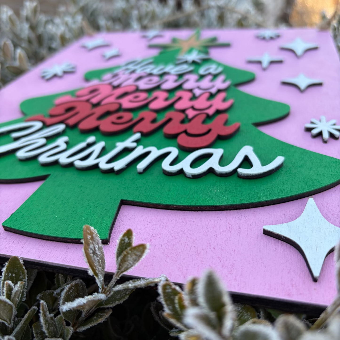Christmas-themed card with a green tree and 'Merry Christmas' text on a pink background, surrounded by plants.