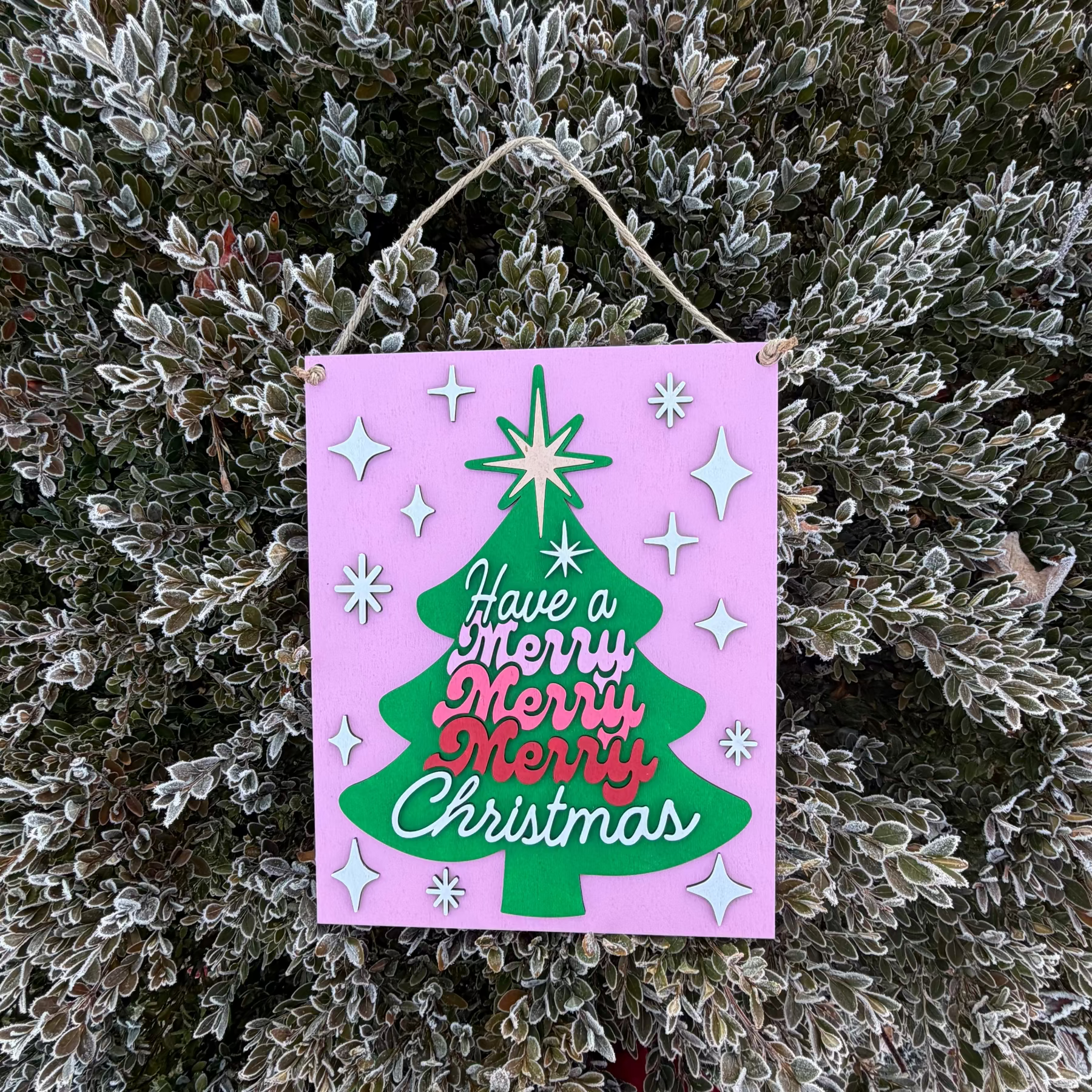 Decorative Christmas sign with a tree design and text, hanging on a frosty bush.