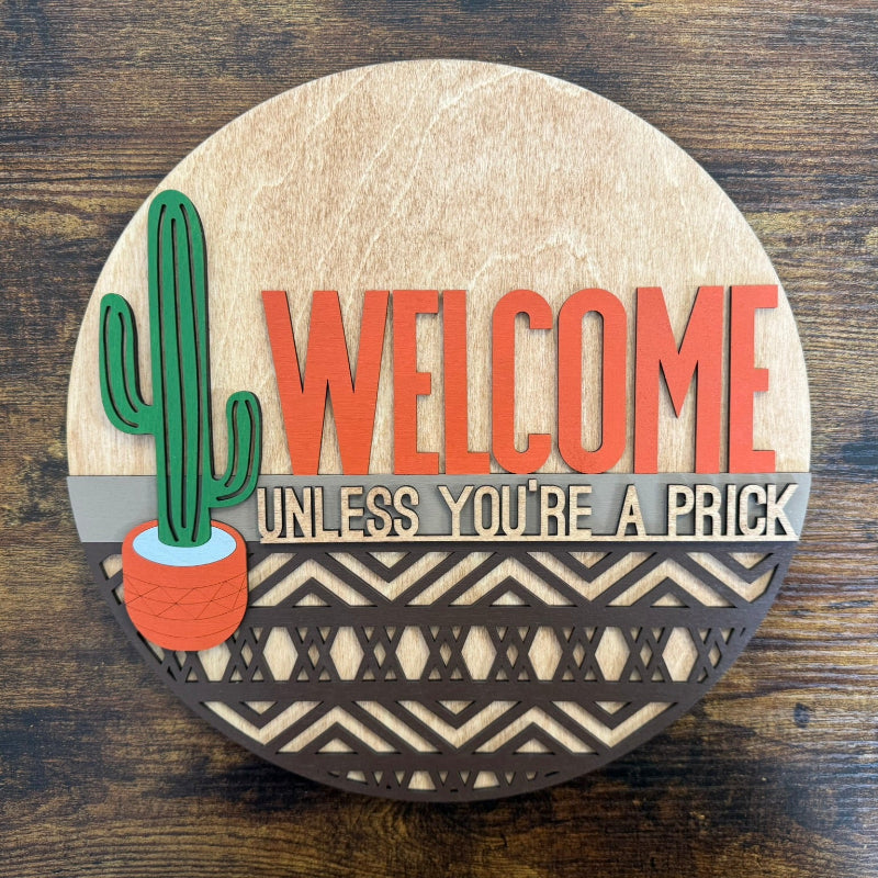 A round decorative plaque with a cactus design and the phrase 'WELCOME UNLESS YOU'RE A PRICK' written on it.